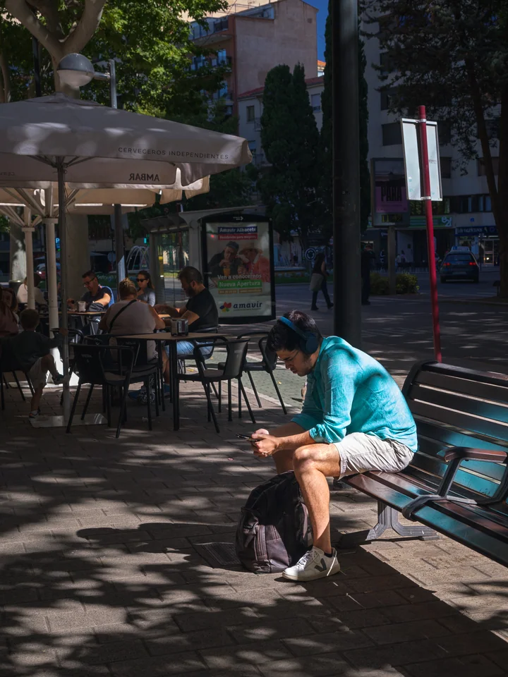 A man with headphones seated on a sunlit bench beside a cafe